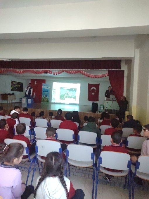 First image shows a classroom with students in red uniforms seated at blue desks, a teacher in a suit pointing at a smartboard displaying content, Turkish flag on wall, educational posters, and children attentively watching. Second image depicts another classroom scene with boys in blue uniforms at orange-trimmed desks, a male teacher in brown jacket gesturing while speaking, world map and flags on walls, backpacks hanging. Third image captures an assembly hall with rows of chairs filled with students in red uniforms, stage with Turkish flags, projector screen showing presentation, adults in suits on stage addressing audience. Fourth image features a stage setup with a man in gray vest speaking at podium, projector displaying text and images related to zero waste including a cartoon character, blue recycling bins nearby, small group of children seated in front rows.