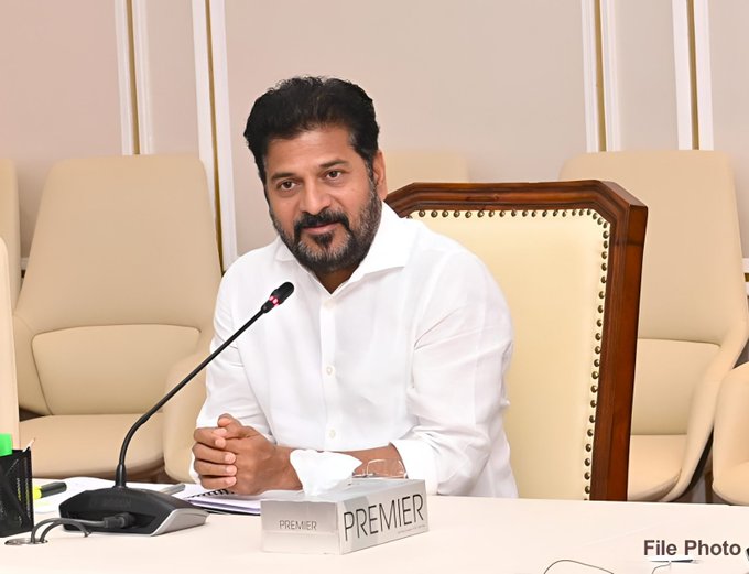 A man with short dark hair and a mustache wearing a white shirt sits at a wooden table in a room with beige walls and wooden accents holding his hands together in front of him facing a microphone on the table with a nameplate reading PREMIER in front of him surrounded by empty beige chairs and a wooden chair to his side with a notebook and pen visible on the table