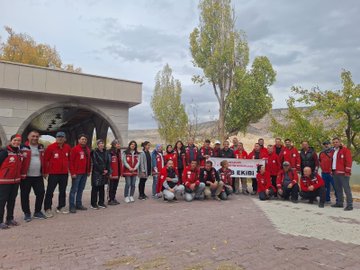 First image shows a large group of people mostly wearing red jackets standing and sitting in front of a stone archway building with poplar trees and rocky hills in the background, holding a banner that reads AKUB. Second image depicts the group walking along a dirt path through autumn foliage with yellow trees and a dirt embankment, including a white dog nearby. Third image captures participants in red jackets setting up a gray and yellow tent near white cars parked on dirt ground surrounded by fall trees and a stone wall. Fourth image features the group in red jackets circled around a campfire on the ground in a wooded area with cars and trees in the background.