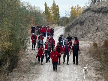 First image shows a large group of people mostly wearing red jackets standing and sitting in front of a stone archway building with poplar trees and rocky hills in the background, holding a banner that reads AKUB. Second image depicts the group walking along a dirt path through autumn foliage with yellow trees and a dirt embankment, including a white dog nearby. Third image captures participants in red jackets setting up a gray and yellow tent near white cars parked on dirt ground surrounded by fall trees and a stone wall. Fourth image features the group in red jackets circled around a campfire on the ground in a wooded area with cars and trees in the background.