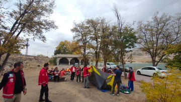 First image shows a large group of people mostly wearing red jackets standing and sitting in front of a stone archway building with poplar trees and rocky hills in the background, holding a banner that reads AKUB. Second image depicts the group walking along a dirt path through autumn foliage with yellow trees and a dirt embankment, including a white dog nearby. Third image captures participants in red jackets setting up a gray and yellow tent near white cars parked on dirt ground surrounded by fall trees and a stone wall. Fourth image features the group in red jackets circled around a campfire on the ground in a wooded area with cars and trees in the background.