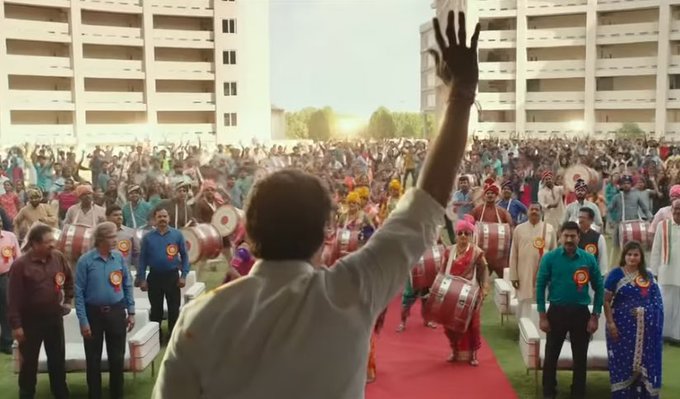 First image shows a large enthusiastic crowd in colorful traditional Indian attire gathered on a green lawn in front of modern white buildings under a clear sky, with several people raising hands in celebration, drummers playing large drums, and a central figure in white shirt and orange scarf standing on a red carpet stage waving hand energetically. Second image depicts an older man with mustache wearing white shirt and shawl sitting in a vintage room with blue curtains, lamp, wooden furniture, and decorative items, looking serious with intense expression.
