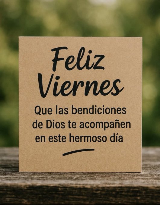 Brown greeting card placed on a wooden surface with a blurred green outdoor background visible behind it. The card displays bold black text reading Feliz Viernes at the top. Below it, the text states Que las bendiciones de Dios te acompañen en este hermoso día, conveying a Friday blessing message.