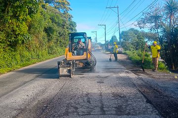 Multiple images show road maintenance scenes with workers in yellow shirts and helmets using shovels to handle asphalt on a roadside surrounded by green vegetation and utility poles. A yellow loader excavates damaged pavement, while another machine spreads asphalt. Traffic cones and a white truck are present, with clear skies overhead. Workers pour and level black asphalt material on the road surface. No watermarks visible.