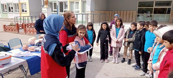 First image shows group of children and women including some wearing headscarves gathered around table with orange bag in school courtyard with buildings and statue in background. Second image depicts instructor in red jacket holding blue ribbon demonstrating to children around table with mannequin and medical supplies. Third image features two instructors one in blue headscarf and one in red jacket showing bandaged arm on boy at blue table with medical props. Fourth image illustrates instructor in red jacket with reflective vest interacting with group of children near table with mannequin and pillows in school yard.
