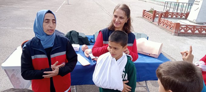 First image shows group of children and women including some wearing headscarves gathered around table with orange bag in school courtyard with buildings and statue in background. Second image depicts instructor in red jacket holding blue ribbon demonstrating to children around table with mannequin and medical supplies. Third image features two instructors one in blue headscarf and one in red jacket showing bandaged arm on boy at blue table with medical props. Fourth image illustrates instructor in red jacket with reflective vest interacting with group of children near table with mannequin and pillows in school yard.
