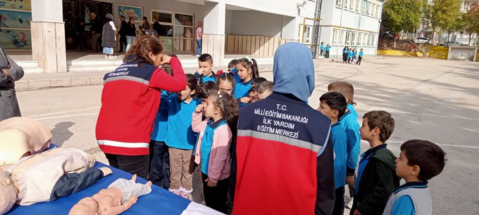 First image shows group of children and women including some wearing headscarves gathered around table with orange bag in school courtyard with buildings and statue in background. Second image depicts instructor in red jacket holding blue ribbon demonstrating to children around table with mannequin and medical supplies. Third image features two instructors one in blue headscarf and one in red jacket showing bandaged arm on boy at blue table with medical props. Fourth image illustrates instructor in red jacket with reflective vest interacting with group of children near table with mannequin and pillows in school yard.