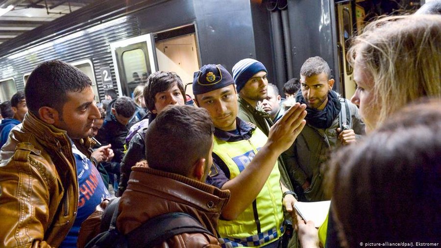 Crowded subway platform scene with multiple people including men in winter jackets and hats gathered around a police officer in yellow vest and blue cap who is gesturing with one hand extended while holding papers in the other. Diverse group of individuals some with backpacks appear engaged in conversation or interaction near train doors and graffiti-covered walls. Officer stands out with uniform and badge visible amid the cluster of onlookers.
