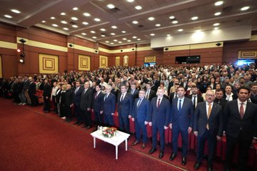 First image shows a man in a dark suit with a red tie standing at a white podium with microphones including one marked with a red logo, speaking towards the camera, with a large backdrop featuring the Turkish flag and a portrait of Recep Tayyip Erdoğan, blue panels on sides, and a red star element. Second image depicts a large group of about 50-60 people mostly men in dark suits and some women in formal attire standing in rows on a red carpeted area in a warmly lit hall with beige walls and geometric patterns, a small white table with flowers in front, and overhead lights. Third image captures a man in a dark suit with mustache sitting and holding a young girl in white top on his lap, another girl in pink headband and white shirt standing beside smiling, red seats around, flowers on a table nearby, and audience blurred in background. Fourth image shows a wide group of around 40 diverse participants including men and women in various formal outfits standing on a stage with red curtains, a central banner with Turkish emblem and text, flanked by large Turkish flags and portraits of Recep Tayyip Erdoğan, wooden floor, and a small table with flowers below.