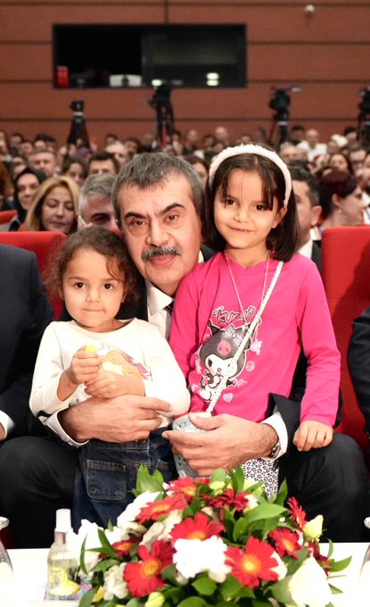 First image shows a man in a dark suit with a red tie standing at a white podium with microphones including one marked with a red logo, speaking towards the camera, with a large backdrop featuring the Turkish flag and a portrait of Recep Tayyip Erdoğan, blue panels on sides, and a red star element. Second image depicts a large group of about 50-60 people mostly men in dark suits and some women in formal attire standing in rows on a red carpeted area in a warmly lit hall with beige walls and geometric patterns, a small white table with flowers in front, and overhead lights. Third image captures a man in a dark suit with mustache sitting and holding a young girl in white top on his lap, another girl in pink headband and white shirt standing beside smiling, red seats around, flowers on a table nearby, and audience blurred in background. Fourth image shows a wide group of around 40 diverse participants including men and women in various formal outfits standing on a stage with red curtains, a central banner with Turkish emblem and text, flanked by large Turkish flags and portraits of Recep Tayyip Erdoğan, wooden floor, and a small table with flowers below.