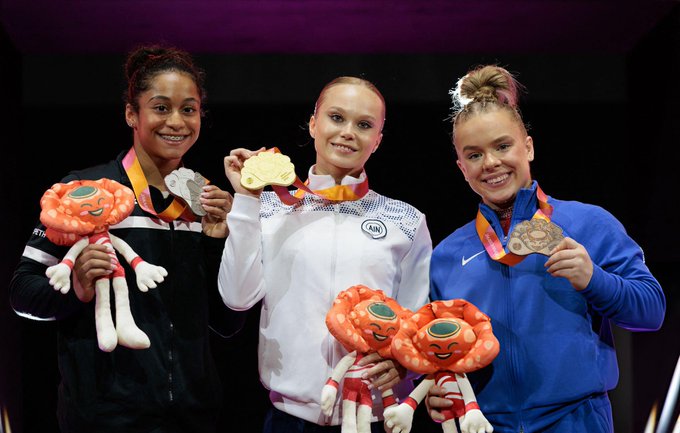 Three female gymnasts stand on a podium after the womens vault event holding their medals and plush toys shaped like orange flowers. The central athlete wears a white jacket with a blue emblem and holds a gold medal around her neck smiling broadly. The left athlete in a black top with orange accents holds a silver medal and a plush toy. The right athlete in a blue top holds a bronze medal and two plush toys. The background is dark with purple lighting.