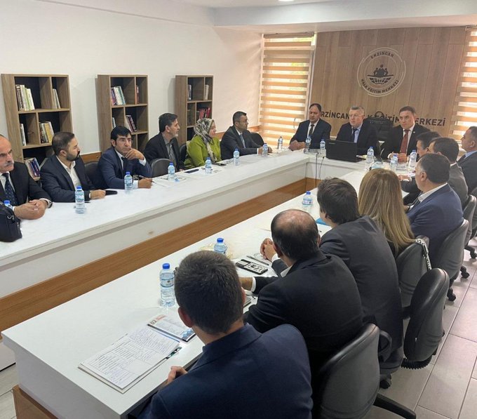 First image shows a large group of about 20 people standing in a row on a paved area outside a building entrance with a sign reading ERZİNCAN HALK EĞİTİMİ MERKEZİ above the door, individuals dressed in formal suits and some in traditional attire holding documents, cloudy sky in background with trees. Second image depicts a group of around 15 people seated around an oval white table in a meeting room with bookshelves and a wall banner reading ERZİNCAN HALK EĞİTİMİ MERKEZİ MÜDÜRLÜĞÜ, participants in business suits some wearing headscarves, water bottles and documents on the table, laptops and microphones present.