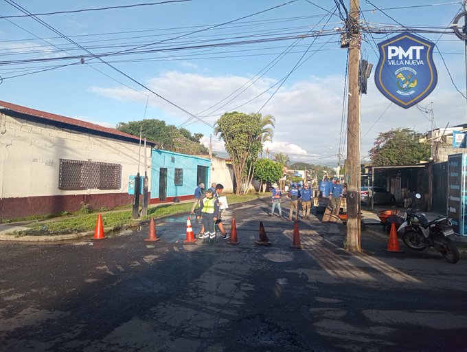 Street scene in urban area with several police officers in blue uniforms and yellow safety vests standing near orange traffic cones on asphalt road. Motorcycle parked nearby. Buildings with blue and red walls in background. Palm trees and utility poles visible. Blue and white shield sign with PMT VILLA NUEVA text on pole.