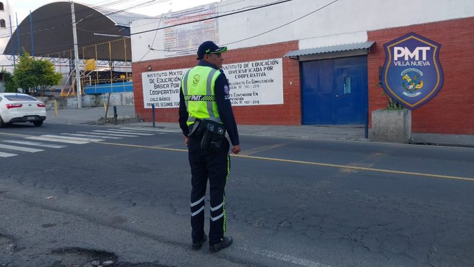 A traffic police officer in a yellow high-visibility vest and dark pants with reflective stripes stands facing away at a crosswalk on a street, gesturing with one arm extended. A white sedan is parked nearby on the road. Behind the officer is a red brick wall with a blue door, a PMT VILLANUEVA shield emblem, and white posters reading Educacion Vial and other text. Construction scaffolding and a large arched structure are visible in the background under a clear sky.