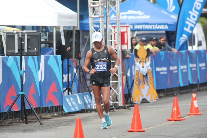 A male athlete wearing a white cap, black sleeveless top with Guatemala logo, number 232 bib, and blue shorts runs on a street during a race event. Blue barriers and orange cones line the path. Spectators in yellow shirts stand behind barriers. Blue tents with Hidrandina and Instantanea logos are visible in the background along with speakers and a yellow shark mascot figure.