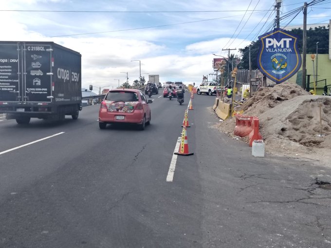 A busy urban road scene with a red car driving in the left lane, several orange traffic cones marking the closed right lane, a white truck parked nearby, construction barriers and dirt piles indicating ongoing works, a large blue sign reading P.M.T. VILLA NUEVA on the roadside, and overhead power lines and buildings in the background under a partly cloudy sky.