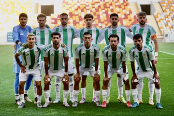 Scoreboard displays Malatya Yeşilyurtspor logo on left with 0, Kahramanmaraş SK logo on right with 2, text Mac Sonucu below. Group of players in green and white striped jerseys stand on field facing camera. Officials in red jerseys and black shorts pose with referee in center holding whistle, soccer ball at feet. Fans including woman in headscarf, man, and child hold Malatya Yeşilyurtspor scarf in stadium seats with orange and yellow surroundings.