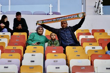 Scoreboard displays Malatya Yeşilyurtspor logo on left with 0, Kahramanmaraş SK logo on right with 2, text Mac Sonucu below. Group of players in green and white striped jerseys stand on field facing camera. Officials in red jerseys and black shorts pose with referee in center holding whistle, soccer ball at feet. Fans including woman in headscarf, man, and child hold Malatya Yeşilyurtspor scarf in stadium seats with orange and yellow surroundings.