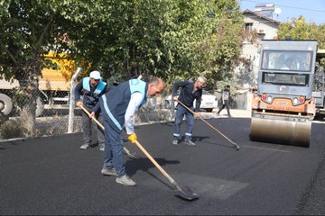 First image shows three workers in blue uniforms and vests using long-handled brooms to smooth fresh black asphalt on a road surface, with a yellow truck and orange roller machine nearby, surrounded by a chain-link fence, trees, and parked cars in a residential area. Second image depicts a long straight newly paved black asphalt road stretching through a rural landscape lined with green trees, yellow bushes, fields, and distant buildings under a clear sky. Third image is an overhead view of construction site with workers in blue uniforms spreading asphalt from a green truck with yellow canopy, near houses, trees, and a white car, with scaffolding and dumpsters present. Fourth image is a wider aerial perspective of a freshly paved asphalt road running through agricultural fields with scattered houses, tents, and vegetation on both sides under sunny conditions.