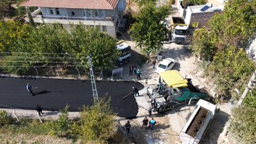 First image shows three workers in blue uniforms and vests using long-handled brooms to smooth fresh black asphalt on a road surface, with a yellow truck and orange roller machine nearby, surrounded by a chain-link fence, trees, and parked cars in a residential area. Second image depicts a long straight newly paved black asphalt road stretching through a rural landscape lined with green trees, yellow bushes, fields, and distant buildings under a clear sky. Third image is an overhead view of construction site with workers in blue uniforms spreading asphalt from a green truck with yellow canopy, near houses, trees, and a white car, with scaffolding and dumpsters present. Fourth image is a wider aerial perspective of a freshly paved asphalt road running through agricultural fields with scattered houses, tents, and vegetation on both sides under sunny conditions.