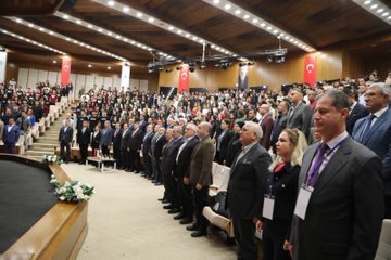 First image shows a man in a dark suit standing at a podium speaking into a microphone with a large projected portrait of an elderly man wearing glasses and a red scarf on a screen behind him flanked by banners reading Vefatının 50 Yılında Arif Nihat Asya Sempozyumu and another banner with the mans portrait. Second image depicts another man in a suit at a podium with a similar large projected portrait of the elderly man on the screen and a banner titled Vefatının 50 Yılında Arif Nihat Asya Sempozyumu. Third image captures a large audience of people mostly in formal attire standing and applauding in a spacious auditorium with wooden paneling multiple Turkish flags on stage and floral decorations. Fourth image illustrates a crowded tiered auditorium filled with seated attendees in business attire during a conference with stage setup including a podium red carpet and banners along with Turkish flags.