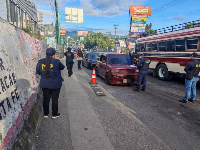 Several police officers in dark blue uniforms with yellow vests stand on a street near a red compact car positioned between orange traffic cones. A red and white bus is parked nearby behind a concrete barrier with graffiti reading parts of words like MACAN and FETA. In the background, a gray van with open doors, billboards advertising Sumar and other signs, a wall with colorful graffiti, and distant green hills under a partly cloudy sky are visible.