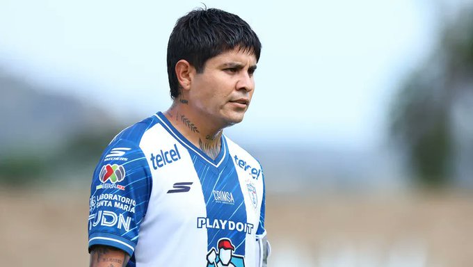 A man with short dark hair and a serious expression stands outdoors on a grassy field under a clear blue sky. He wears a blue and white Pachuca soccer jersey with sponsor patches including Telcel, Telmex, and Play, along with a neck tattoo visible. The jersey has the teams logo and is designed for professional play.