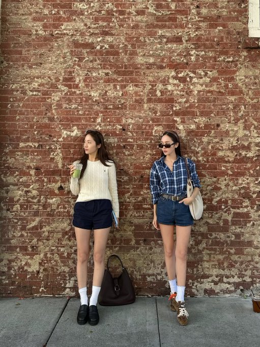 First image shows two women standing against a red brick wall one wearing a white sweater and dark shorts holding a green drink the other in a blue plaid shirt dark shorts and sunglasses holding a beige bag both wearing white socks and different shoes with a brown bag on the ground. Second image depicts the same women against the brick wall one holding an apple the other a phone both in similar outfits with white socks and shoes. Third image captures the women walking hand-in-hand on a sidewalk near a Starlet theater sign with autumn trees one in a white sweater dark shorts and loafers the other in a blue plaid shirt dark shorts and sneakers carrying bags small figures visible.