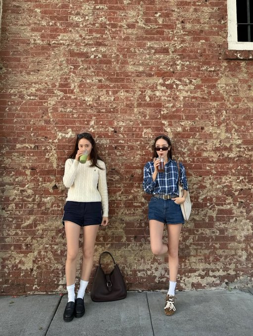 First image shows two women standing against a red brick wall one wearing a white sweater and dark shorts holding a green drink the other in a blue plaid shirt dark shorts and sunglasses holding a beige bag both wearing white socks and different shoes with a brown bag on the ground. Second image depicts the same women against the brick wall one holding an apple the other a phone both in similar outfits with white socks and shoes. Third image captures the women walking hand-in-hand on a sidewalk near a Starlet theater sign with autumn trees one in a white sweater dark shorts and loafers the other in a blue plaid shirt dark shorts and sneakers carrying bags small figures visible.