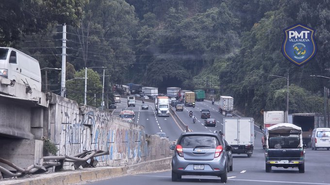 Highway scene with multiple lanes showing heavy traffic congestion including white vans, trucks, cars, and a small gray vehicle in the foreground. Surrounding elements include graffiti-covered concrete barriers, utility poles, trees, and an overpass structure. A blue police badge labeled PMT is prominently displayed in the upper right corner.