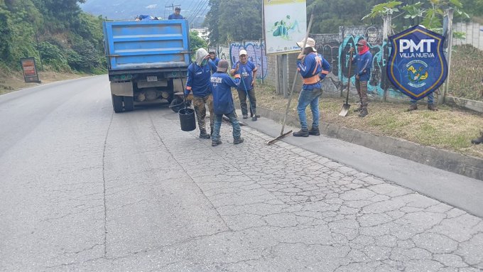 Several uniformed police officers wearing blue shirts with PMT patches and hats stand on a paved roadside next to a large blue dump truck loaded with gravel. They hold brooms and shovels, sweeping and working on the ground near a chain-link fence. A green banner with PMT logo and text hangs on the fence, and lush green hills are visible in the background under a clear sky.