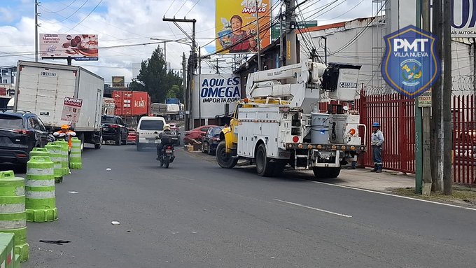 Urban street scene on Calzada Aguilar Batres with green traffic barriers, a white utility truck with bucket lift extended near power lines, parked vehicles including trucks and motorcycles, billboards advertising Omega and other products, a red gate, workers in yellow vests, and a blue shield logo of PMT Villa Nueva on a pole.