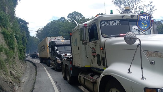 Multiple large trucks including a white tow truck with license plate C-683BP and PMT logo on the side are positioned on a narrow, curved uphill road surrounded by green hills and trees. The tow truck is attached to a yellow trailer truck with mechanical issues. Other trucks are queued behind on the road. The scene appears during daytime with cloudy skies.