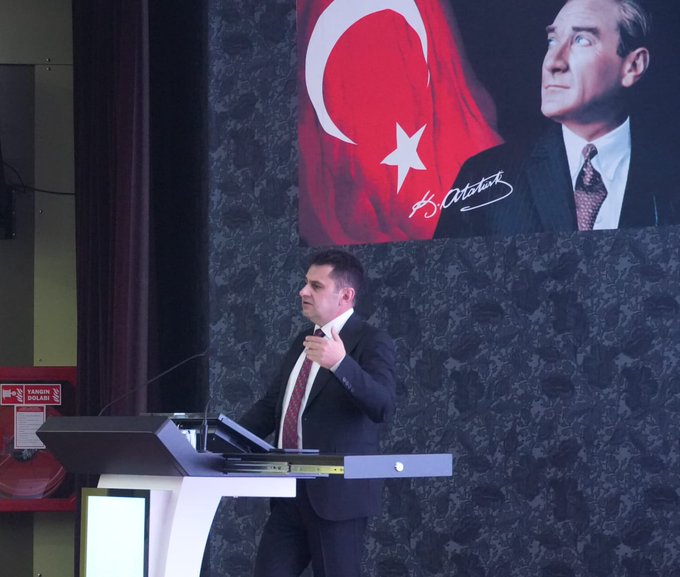 First image shows a man in a dark suit standing at a podium speaking into a microphone, with a large Turkish flag and a portrait of a man with a signature on the wall behind him in an auditorium setting. Second image depicts a large audience of mostly men in suits seated in rows facing a stage where a man in a dark suit speaks at a podium, with Turkish flags and flowers on stage in a conference hall. Third image features a screen displaying text about OGM Management Board meeting details including dates and agenda on vocational education, with a man in suit at podium and audience seated in front.
