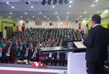 First image shows a man in a dark suit standing at a podium speaking into a microphone, with a large Turkish flag and a portrait of a man with a signature on the wall behind him in an auditorium setting. Second image depicts a large audience of mostly men in suits seated in rows facing a stage where a man in a dark suit speaks at a podium, with Turkish flags and flowers on stage in a conference hall. Third image features a screen displaying text about OGM Management Board meeting details including dates and agenda on vocational education, with a man in suit at podium and audience seated in front.