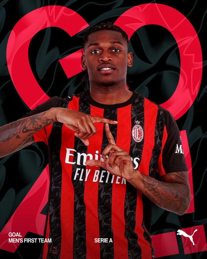 A young Black male soccer player with tattoos on his arms stands pointing both index fingers forward, wearing a black and red striped AC Milan jersey with Puma logo, Emirates Fly Better sponsor text, number 22, and Serie A patch on the sleeve, against a dark background with red heart shapes and text reading GOAL FIRST TEAM.
