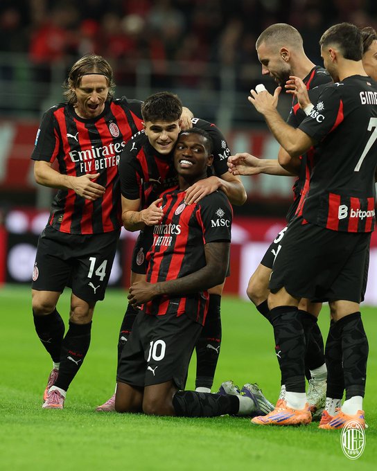 Group of male soccer players in red and black AC Milan jerseys with sponsor logos like Emirates Fly Better and MSC on chests celebrating on green field with stadium seating and crowd blurred in background. Players include one with long hair number 14 kneeling and smiling one with short hair number 10 embracing from behind another with bald head clapping and one with dreadlocks number 7 standing nearby all wearing black shorts and mismatched socks some pink some orange cleats visible.
