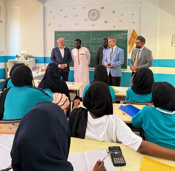 First image shows a classroom with a blackboard displaying math equations and a map, several adults in suits and traditional attire standing and interacting with female students in hijabs seated at desks with notebooks and calculators. Second image depicts a colorful classroom with heart decorations, adults in formal wear surrounding young children in uniforms sitting around a round table painting on large sheets of paper using crayons. Third image features a wall mural labeled Afrika with a map of Africa, an older man in a suit shaking hands with teenage boys in teal uniforms standing by desks with books. Fourth image captures a stage event with a backdrop of the Türkiye Maarif Vakfı logo, children in uniforms holding flags of various countries including Turkish and Romanian, standing in a line with balloons and seated audience including a man in traditional attire.