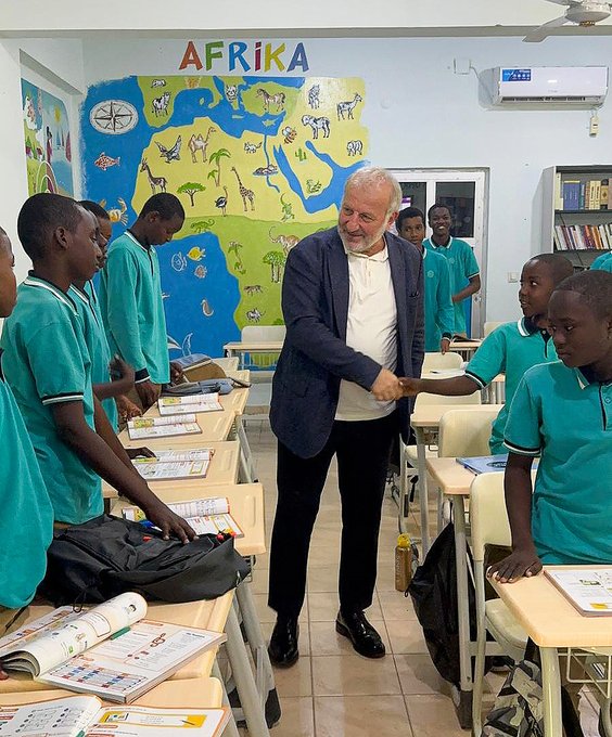 First image shows a classroom with a blackboard displaying math equations and a map, several adults in suits and traditional attire standing and interacting with female students in hijabs seated at desks with notebooks and calculators. Second image depicts a colorful classroom with heart decorations, adults in formal wear surrounding young children in uniforms sitting around a round table painting on large sheets of paper using crayons. Third image features a wall mural labeled Afrika with a map of Africa, an older man in a suit shaking hands with teenage boys in teal uniforms standing by desks with books. Fourth image captures a stage event with a backdrop of the Türkiye Maarif Vakfı logo, children in uniforms holding flags of various countries including Turkish and Romanian, standing in a line with balloons and seated audience including a man in traditional attire.