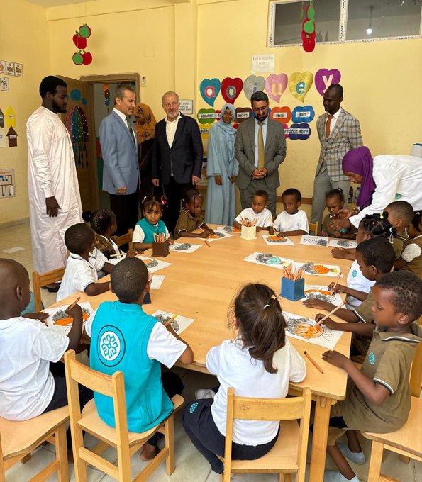 First image shows a classroom with a blackboard displaying math equations and a map, several adults in suits and traditional attire standing and interacting with female students in hijabs seated at desks with notebooks and calculators. Second image depicts a colorful classroom with heart decorations, adults in formal wear surrounding young children in uniforms sitting around a round table painting on large sheets of paper using crayons. Third image features a wall mural labeled Afrika with a map of Africa, an older man in a suit shaking hands with teenage boys in teal uniforms standing by desks with books. Fourth image captures a stage event with a backdrop of the Türkiye Maarif Vakfı logo, children in uniforms holding flags of various countries including Turkish and Romanian, standing in a line with balloons and seated audience including a man in traditional attire.