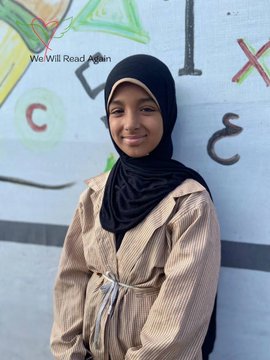 First image shows a young girl with brown skin wearing a black hijab and beige striped long-sleeve shirt sitting on a brown rattan chair holding a purple pen and writing in an open notebook on her lap with Arabic script visible. Background includes a beige tent-like structure and another chair. Overlay text reads We Will Read Again with heart and book emojis. Second image shows the same girl smiling with closed-mouth expression standing in front of a blue wall painted with green heart book logo and We Will Read Again text plus drawings of numbers 1 and letters X C in white chalk and Arabic script.