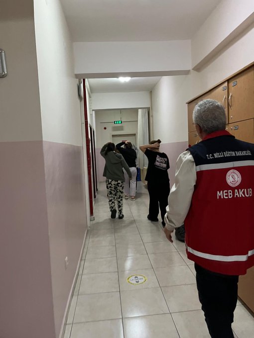 First image shows a hospital or school corridor with light pink and white walls tiled floor several people including a man in red MEB vest standing and walking near wooden lockers green exit sign curtained area and yellow social distancing mark on floor. Second image depicts a dormitory room with two bunk beds wooden frames white window brown clothes bags on beds and floor two people one in red MEB vest kneeling near bed another in black clothing.