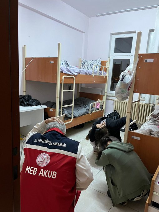 First image shows a hospital or school corridor with light pink and white walls tiled floor several people including a man in red MEB vest standing and walking near wooden lockers green exit sign curtained area and yellow social distancing mark on floor. Second image depicts a dormitory room with two bunk beds wooden frames white window brown clothes bags on beds and floor two people one in red MEB vest kneeling near bed another in black clothing.
