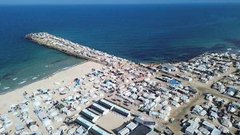 Aerial view shows numerous white tents densely packed along a sandy beach extending to the sea with a pier structure protruding into turquoise water people visible walking near tents and structures scattered across the area under clear skies.