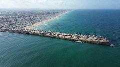 Aerial view shows numerous white tents densely packed along a sandy beach extending to the sea with a pier structure protruding into turquoise water people visible walking near tents and structures scattered across the area under clear skies.