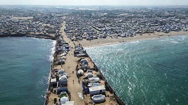 Aerial view shows numerous white tents densely packed along a sandy beach extending to the sea with a pier structure protruding into turquoise water people visible walking near tents and structures scattered across the area under clear skies.