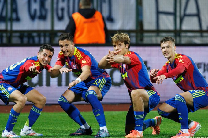 Four male athletes wearing red and blue striped soccer jerseys and blue shorts stand on a green grass field, posing with hands extended forward as if celebrating or signaling. They have short dark hair and athletic builds, positioned side by side in a line. In the background, a man in an orange safety vest stands near a blue stadium banner with white text, under a partly cloudy sky.