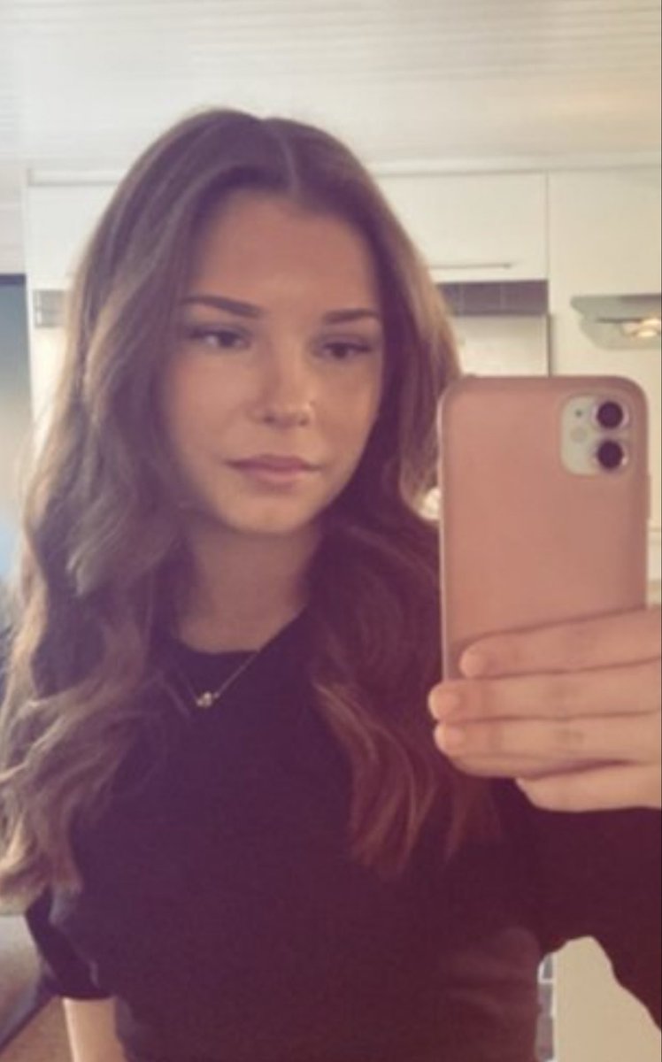 A young woman with long wavy brown hair takes a selfie in a bright indoor room with white walls and wooden cabinets. She wears a black short-sleeved top and a delicate necklace. She holds a pink-cased smartphone close to her face with her right hand showing a neutral expression. The background includes a window with sheer curtains and a shelf.