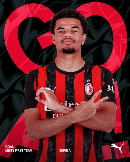 A young male soccer player with curly hair and a beard stands confidently in an AC Milan home jersey featuring red and black vertical stripes with the club crest on the chest and Puma logo, posing with arms crossed and fingers pointing outward in a celebratory gesture, set against a dark background with red heart shapes and text overlays reading GOAL FIRST TEAM SERIE A and M 24.