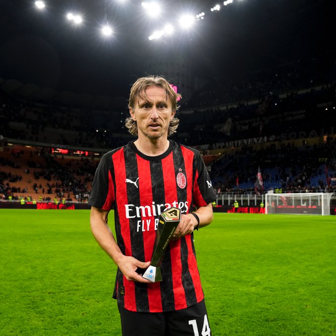 A man with long blond hair wearing a red and black striped AC Milan soccer jersey with number 14 and sponsor logos stands on a green soccer field holding a gold trophy in his right hand. Stadium lights illuminate the scene with a blurred crowd in the stands and a goal net visible in the background. The jersey features Puma branding and Emirates sponsorship.